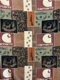 A Carhartt patchwork print 50 x 60” plush blanket. The blanket  features a mix of Realtree camouflage, brown and burgundy Carhartt logos, hunting silhouettes, and vertical orange-brown stripes.