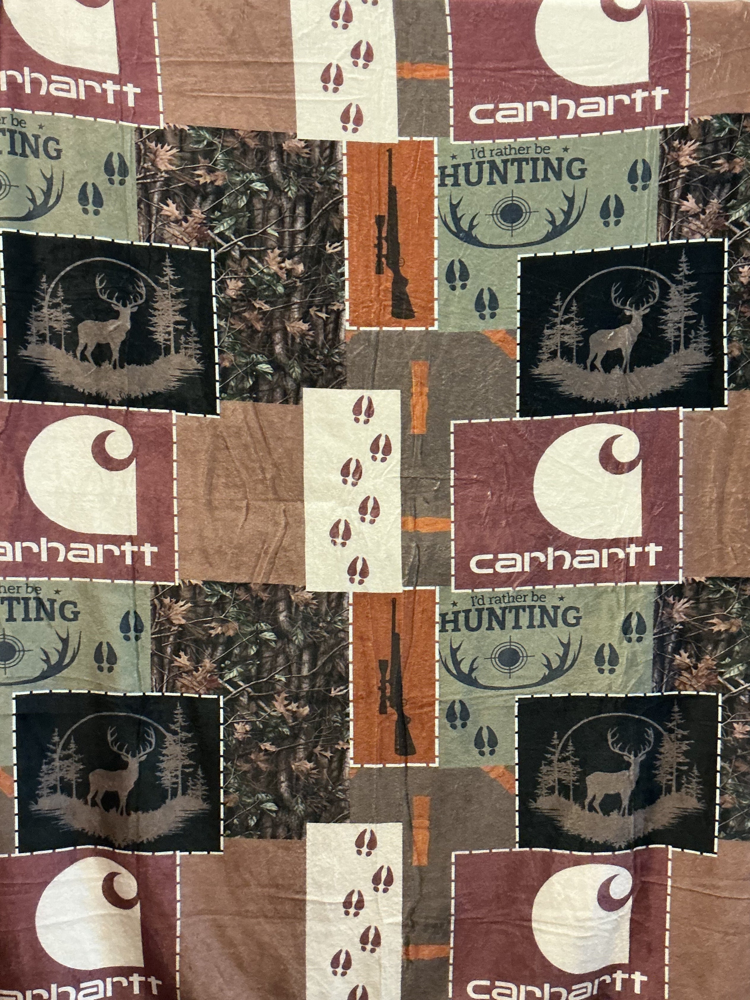 A Carhartt patchwork print 50 x 60” plush blanket. The blanket  features a mix of Realtree camouflage, brown and burgundy Carhartt logos, hunting silhouettes, and vertical orange-brown stripes.