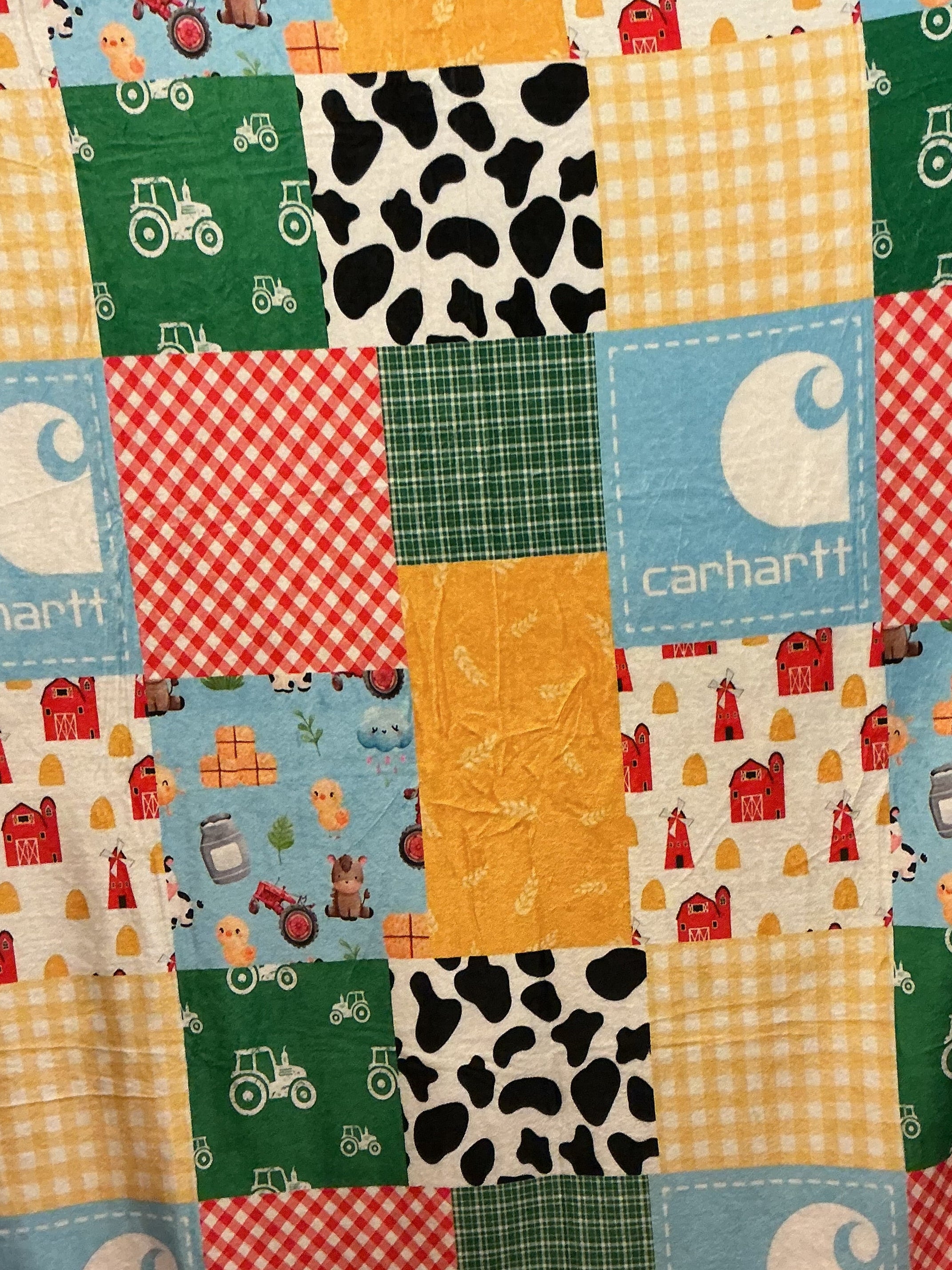 A square patchwork print plush blanket featuring various colorful fabric panels with a Carhartt theme, including the Carhartt logo, tractor prints, cow spots, and gingham patterns.