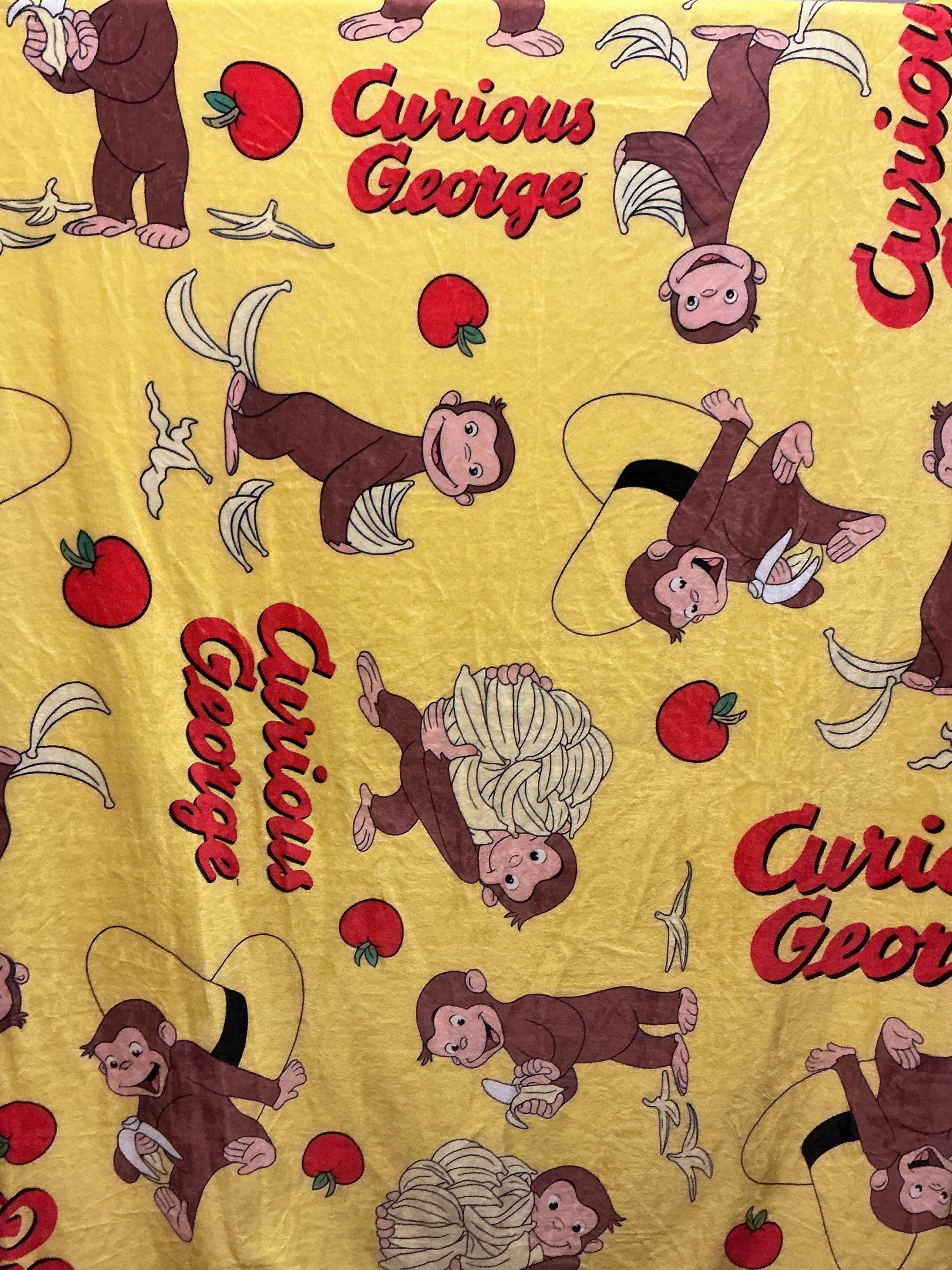 A yellow plush blanket featuring a repeating pattern of Curious George illustrations, red apples, and red 'Curious George' text.