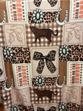 A close-up of a western-themed fabric print featuring cowboys, horseshoes, leopard print, plaid, paw prints, and patches with the words 'DIBS ON THE COWBOY”
