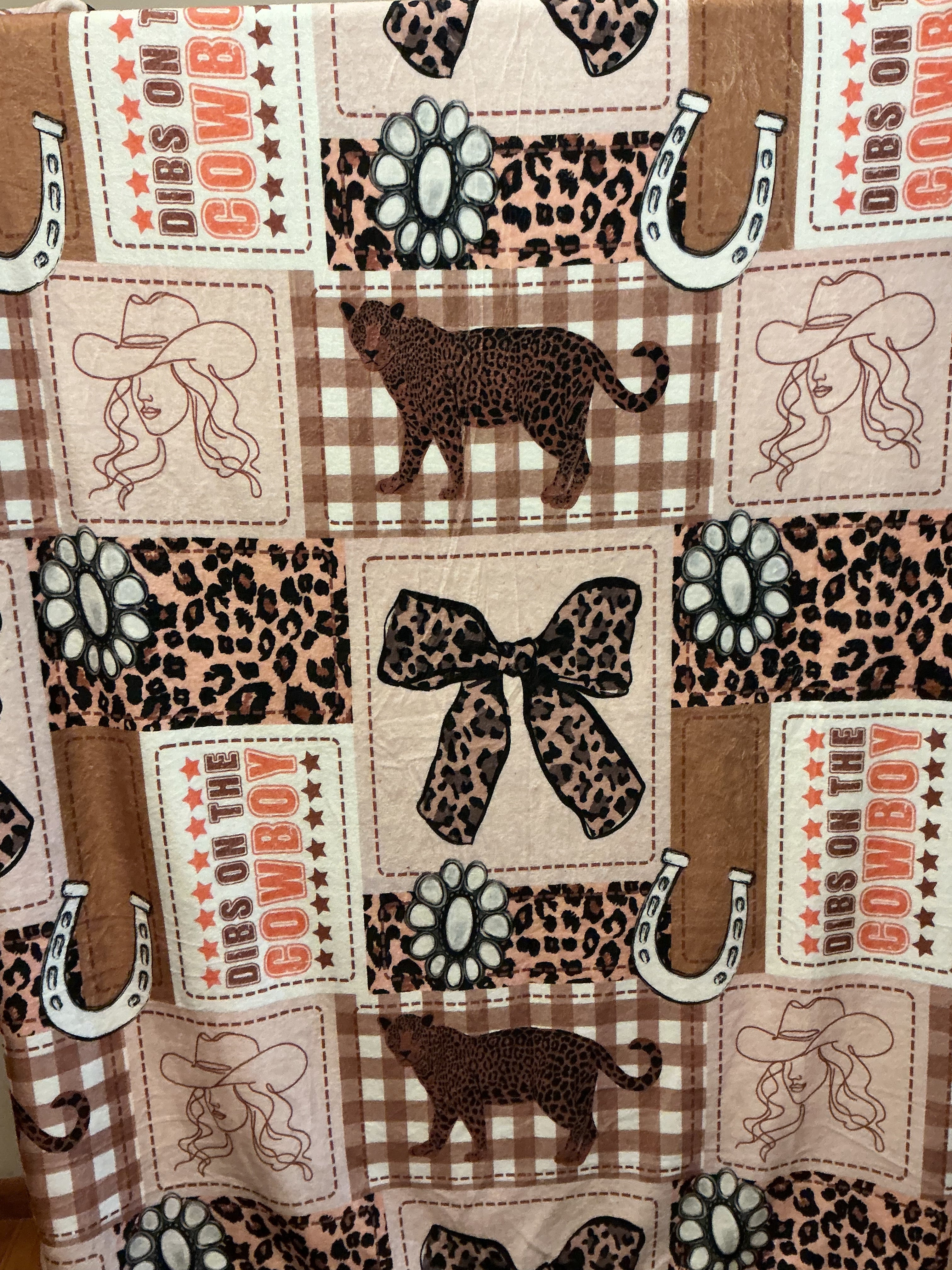 A close-up of a western-themed fabric print featuring cowboys, horseshoes, leopard print, plaid, paw prints, and patches with the words 'DIBS ON THE COWBOY”