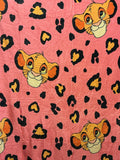 Pink fabric with an allover print of Simba's cartoon faces and black leopard spots.